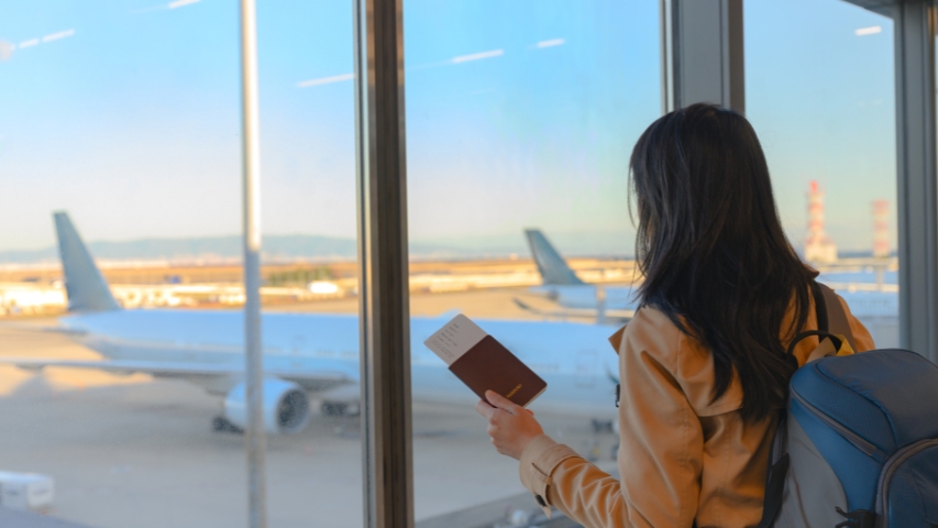 Fotografía de una alumna con un pasaporte en la mano mirando los aviones a través de una cristalera del aeropuerto