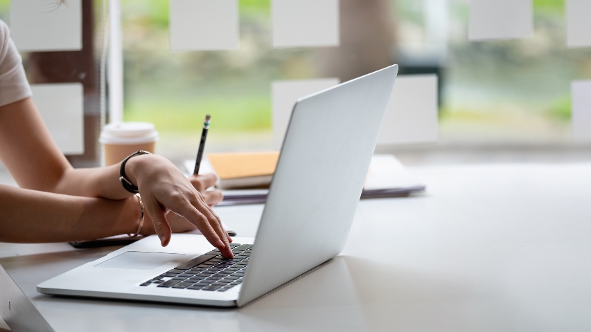 Fotografía de unas manos cruzadas sobre un ordenador, una mano sujeta un lápiz, la otra mano está sobre el teclado